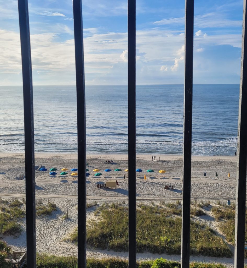 View of the beach with colorful umbrellas, gentle waves, and people walking along the shore, framed by vertical black railings.